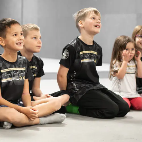 Kinder sitzen in KampfKunstLive-Shirts auf der Matte und hören aufmerksam zu